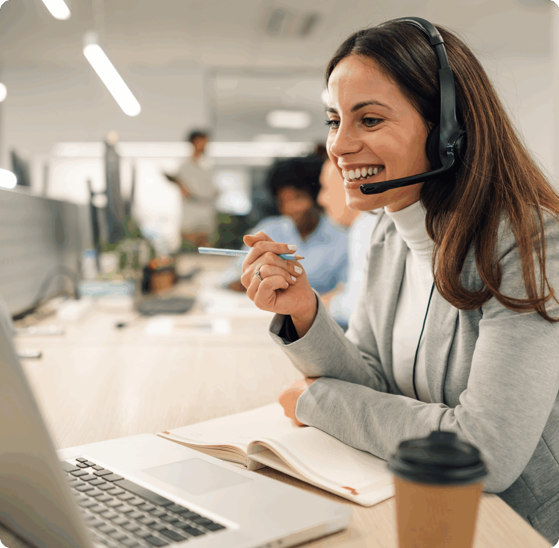 A woman wearing a headset smiling while working on a laptop at a desk in a bright office environment.