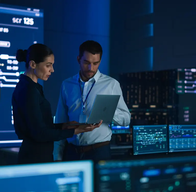 Two IT professionals analyzing data on a laptop in a dimly lit server room filled with monitors and equipment.